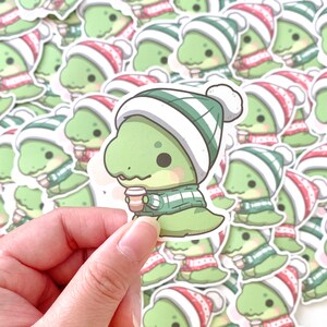 May include: A sheet of stickers featuring a cartoon dinosaur wearing a green and white striped scarf and a red and white Santa hat. The dinosaur is holding a cup of coffee.