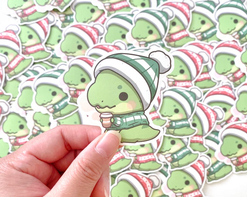 May include: A sheet of stickers featuring a cartoon dinosaur wearing a green and white striped scarf and a red and white Santa hat. The dinosaur is holding a cup of coffee.