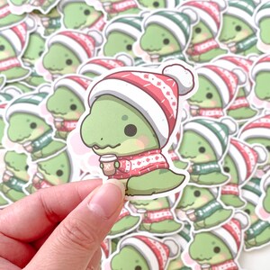 May include: A collection of green dinosaur stickers with red and white winter hats. Each dinosaur is holding a cup of coffee. The stickers are on a white background.
