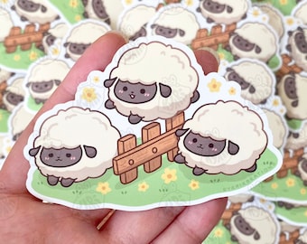 May include: Three white sheep with black faces and legs are standing in a grassy field with yellow flowers. The sheep are near a wooden fence. The sticker is on a hand.