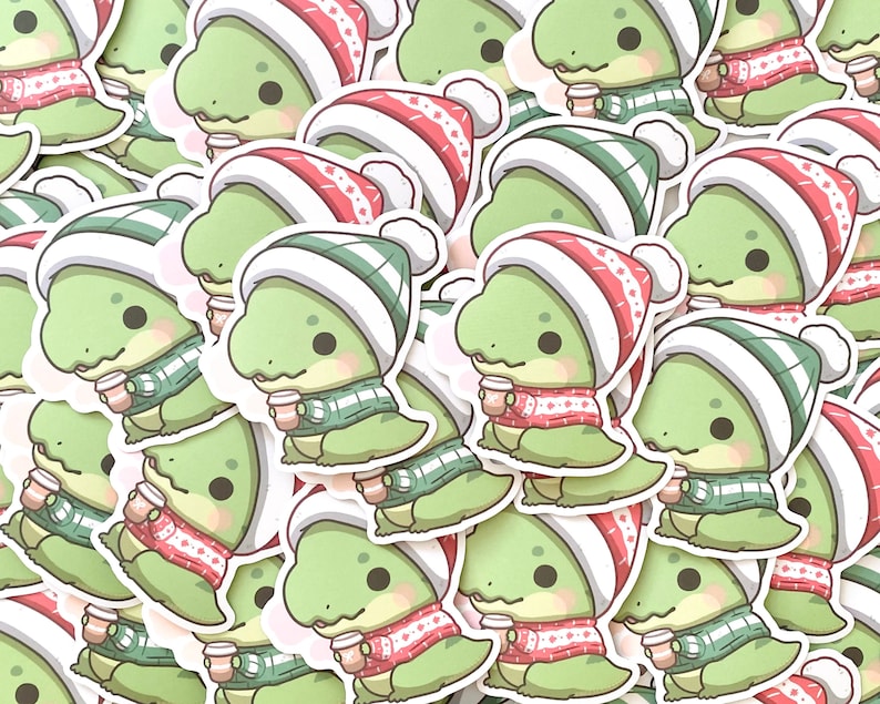 May include: A collection of green dinosaur stickers wearing red and green winter hats and scarves, holding a cup of hot cocoa. The stickers are perfect for adding a festive touch to any project.
