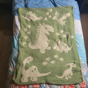 May include: A green crocheted blanket with a white dinosaur design. The blanket features a large dinosaur in the center, surrounded by smaller dinosaurs and footprints.