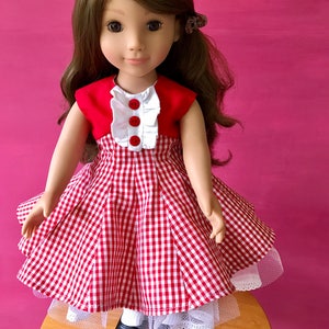 May include: A doll wearing a red and white gingham dress with a white ruffle collar and red buttons. The dress has a white petticoat underneath. The doll has brown hair and is wearing black shoes.
