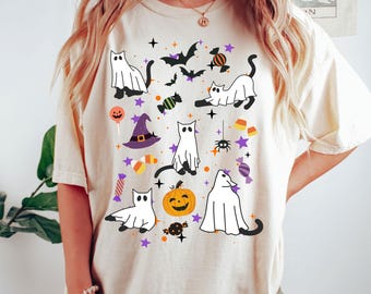 May include: Cream-colored t-shirt featuring a Halloween-themed pattern. The design includes white ghost cats, bats, a jack-o'-lantern, candy, a witch's hat, and stars in orange, purple, and black. The shirt has a relaxed fit.