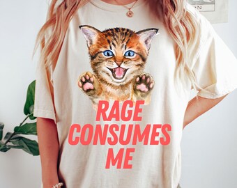 Puede incluir: Una camiseta de color crema con una imagen de dibujos animados de un gato atigrado marrón y naranja con ojos azules. El gato está sonriendo y tiene las patas levantadas. El texto "Rage Consumes Me" está impreso en rojo debajo del gato.