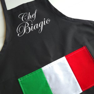 以下が含まれることがあります： 白い筆記体で「Chef Biagio」と書かれた黒いエプロン。緑、白、赤のストライプが入ったイタリア国旗のパッチが縫い付けられています。エプロンには調節可能なネックストラップが付いています。