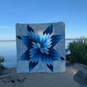 Op de afbeelding: Een grote, vierkante quilt met een geometrisch sterontwerp in blauwtinten. De quilt wordt buiten tentoongesteld, met een meer en blauwe lucht op de achtergrond. Het sterpatroon bestaat uit verschillende tinten blauw.