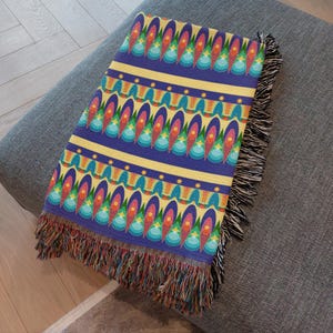 Könnte beinhalten: Eine gefaltete Decke mit einem lebendigen, geometrischen Muster. Das Design zeigt sich wiederholende ovale Formen in Rot-, Orange-, Blau- und Grüntönen auf blauem Hintergrund. Gelbe Streifen und Fransenränder vervollständigen das Design.