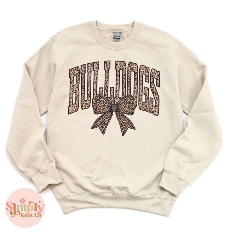 May include: A beige sweatshirt with the word "BULLDOGS" in a leopard print font, arched above a leopard print bow. The sweatshirt has a crew neck and long sleeves.