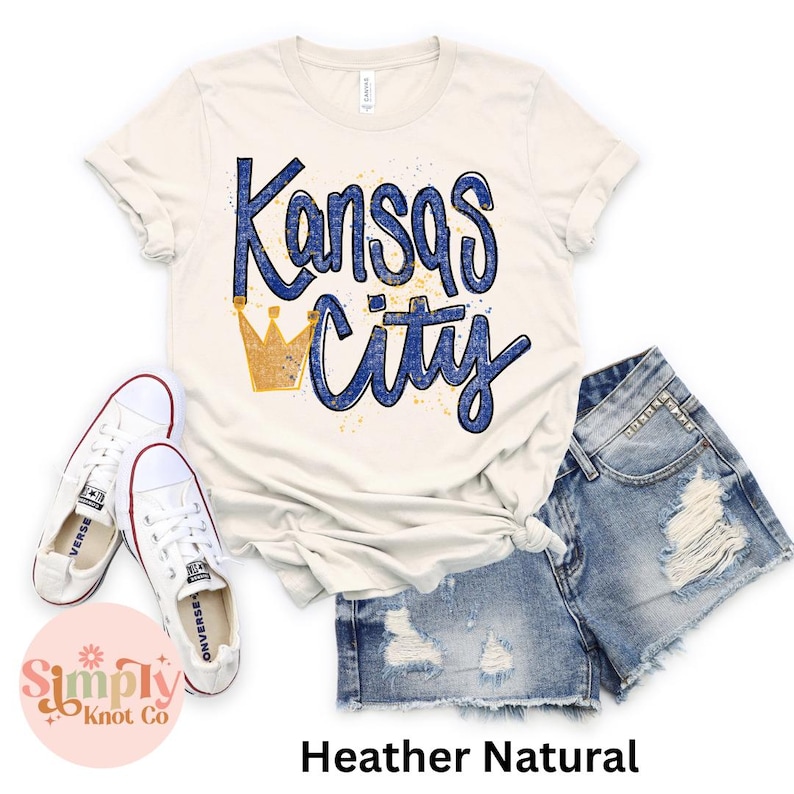 May include: A cream-colored t-shirt with the words "Kansas City" in blue script and a gold crown graphic. The shirt is paired with denim shorts and white sneakers. The text "Heather Natural" is at the bottom.