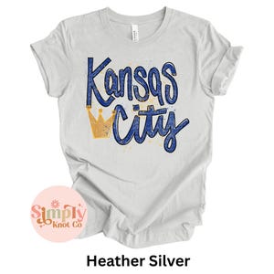 May include: Heather silver t-shirt with the words "Kansas City" in blue script and a gold crown graphic. The shirt has rolled-up sleeves and a tag that says "Canvas". The shirt is a light gray color.