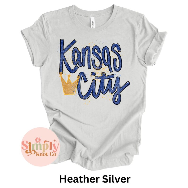 May include: Heather silver t-shirt with the words "Kansas City" in blue script and a gold crown graphic. The shirt has rolled-up sleeves and a tag that says "Canvas". The shirt is a light gray color.