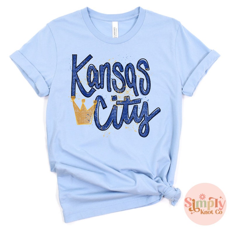 May include: Light blue t-shirt with the words "Kansas City" in blue script and a gold crown graphic. The shirt is made of a soft material and has rolled-up sleeves. The shirt is knotted at the bottom.