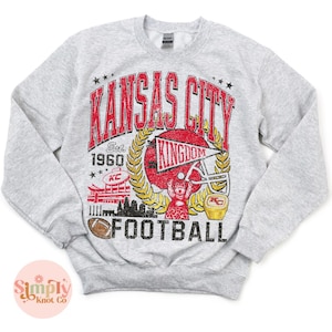 Puede incluir: Sudadera gris claro con un dise&ntilde;o gr&aacute;fico desgastado. El dise&ntilde;o presenta las palabras "Kansas City" en rojo, un casco de f&uacute;tbol y la palabra "Football". La sudadera tiene cuello redondo y mangas largas.
