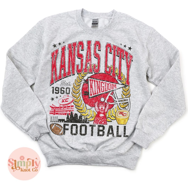 Puede incluir: Sudadera gris claro con un dise&ntilde;o gr&aacute;fico desgastado. El dise&ntilde;o presenta las palabras "Kansas City" en rojo, un casco de f&uacute;tbol y la palabra "Football". La sudadera tiene cuello redondo y mangas largas.