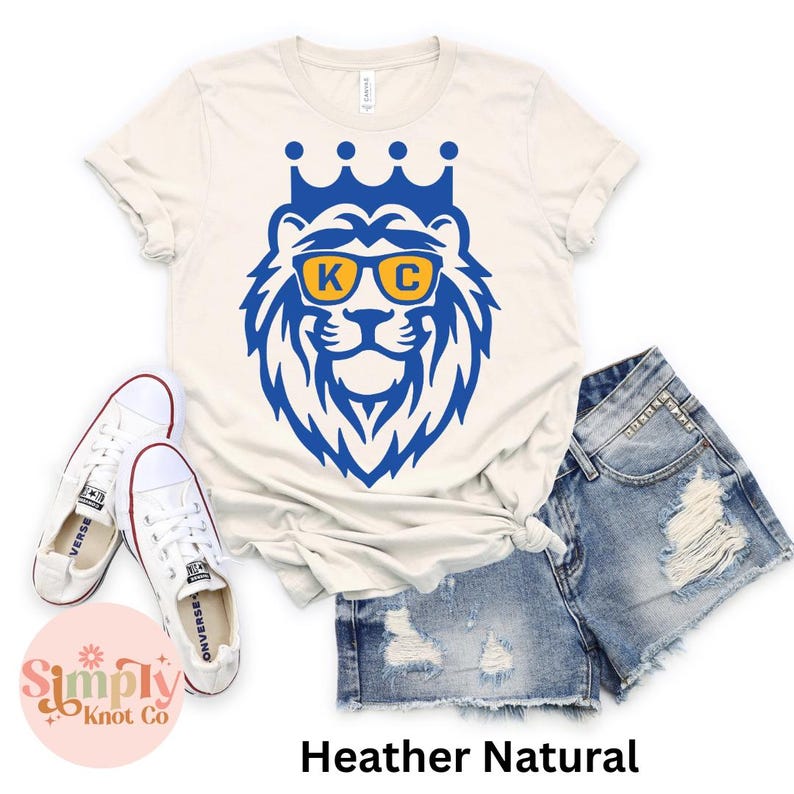 Pu&ograve; includere: Una t-shirt color heather natural con una grafica di leone blu che indossa una corona e occhiali da sole gialli con le lettere K e C sulle lenti. La maglietta &egrave; mostrata insieme a un paio di scarpe da ginnastica bianche e pantaloncini di jeans.