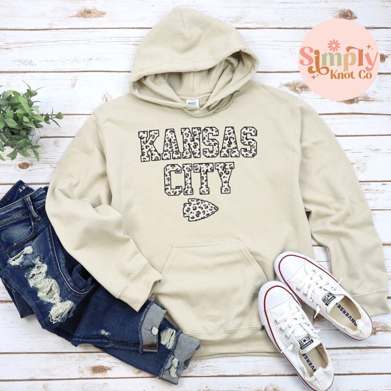 May include: A beige hoodie with a leopard print "Kansas City" graphic and a leopard print arrow on the front. The hoodie has a drawstring hood and a kangaroo pocket.