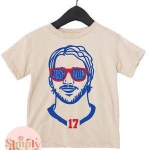 May include: Tan short-sleeve t-shirt featuring a blue and red graphic of a person wearing sunglasses with the text "JOSH ALLEN" on the sunglasses. The number "17" is below the graphic. The shirt is hanging on a black hanger.
