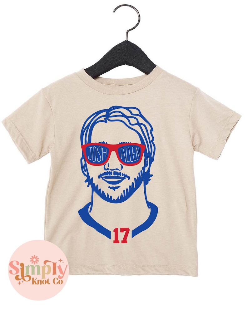 May include: Tan short-sleeve t-shirt featuring a blue and red graphic of a person wearing sunglasses with the text "JOSH ALLEN" on the sunglasses. The number "17" is below the graphic. The shirt is hanging on a black hanger.
