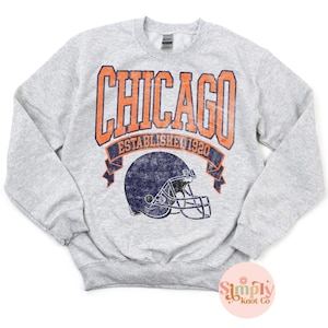 May include: A light gray sweatshirt with the word "CHICAGO" in orange block letters above a navy blue football helmet graphic. The text "ESTABLISHED 1920" is below the word "CHICAGO". The sweatshirt has long sleeves and a crew neck.