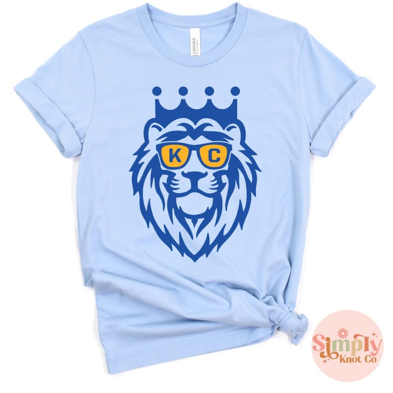 Pu&ograve; includere: Una t-shirt a maniche corte color azzurro con la grafica di un leone che indossa una corona e occhiali da sole gialli con le lettere K e C stampate sulle lenti. La grafica del leone &egrave; realizzata in un colore blu acceso, centrata sul petto.
