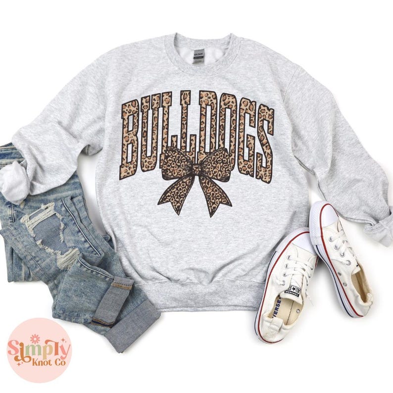 May include: A light gray sweatshirt with the word "BULLDOGS" in a leopard print font and a matching bow graphic. The sweatshirt is paired with blue jeans and white sneakers.