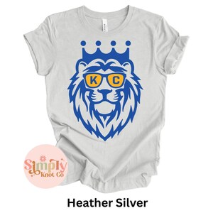 Pu&ograve; includere: Una t-shirt a maniche corte color grigio melange con una grafica blu di un leone che indossa una corona e occhiali da sole gialli con le lettere K e C stampate sulle lenti. Il disegno &egrave; centrato sul petto della t-shirt con scollo rotondo.