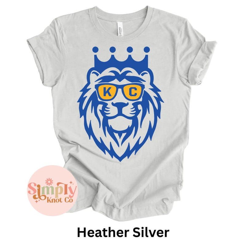 Pu&ograve; includere: Una t-shirt a maniche corte color grigio melange con una grafica blu di un leone che indossa una corona e occhiali da sole gialli con le lettere K e C stampate sulle lenti. Il disegno &egrave; centrato sul petto della t-shirt con scollo rotondo.