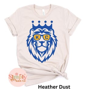 Pu&ograve; includere: Una t-shirt color heather dust con una grafica blu di un leone che indossa una corona e occhiali da sole gialli con le lettere K e C stampate sulle lenti. La maglietta ha maniche corte e un girocollo.