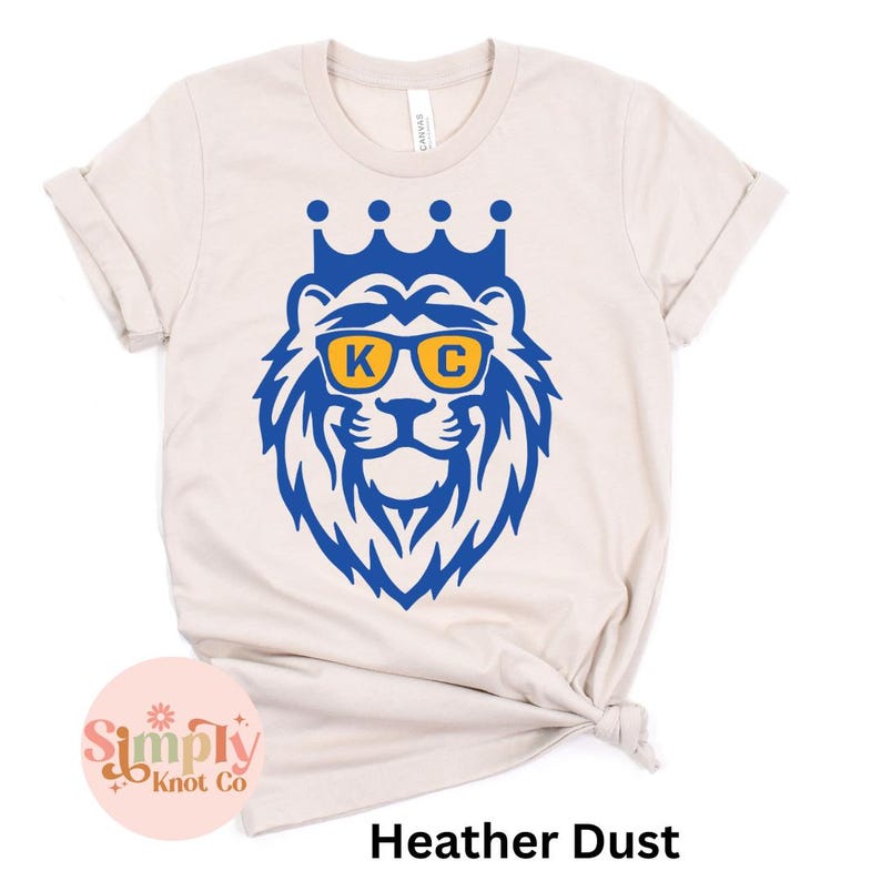 Pu&ograve; includere: Una t-shirt color heather dust con una grafica blu di un leone che indossa una corona e occhiali da sole gialli con le lettere K e C stampate sulle lenti. La maglietta ha maniche corte e un girocollo.
