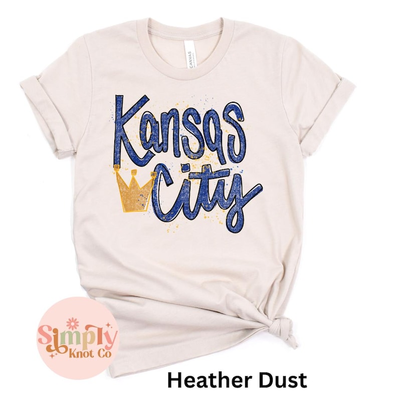 May include: A cream-colored t-shirt with the words "Kansas City" in blue script and a gold crown graphic. The shirt has rolled-up sleeves and a knot at the bottom. The text "Heather Dust" is at the bottom.
