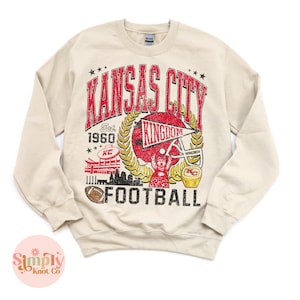 Puede incluir: Sudadera beige con un dise&ntilde;o gr&aacute;fico desgastado. El dise&ntilde;o presenta las palabras "Kansas City Football" en rojo, con un casco de f&uacute;tbol, un bal&oacute;n de f&uacute;tbol y el texto "Est. 1960". La sudadera tiene cuello redondo y mangas largas.