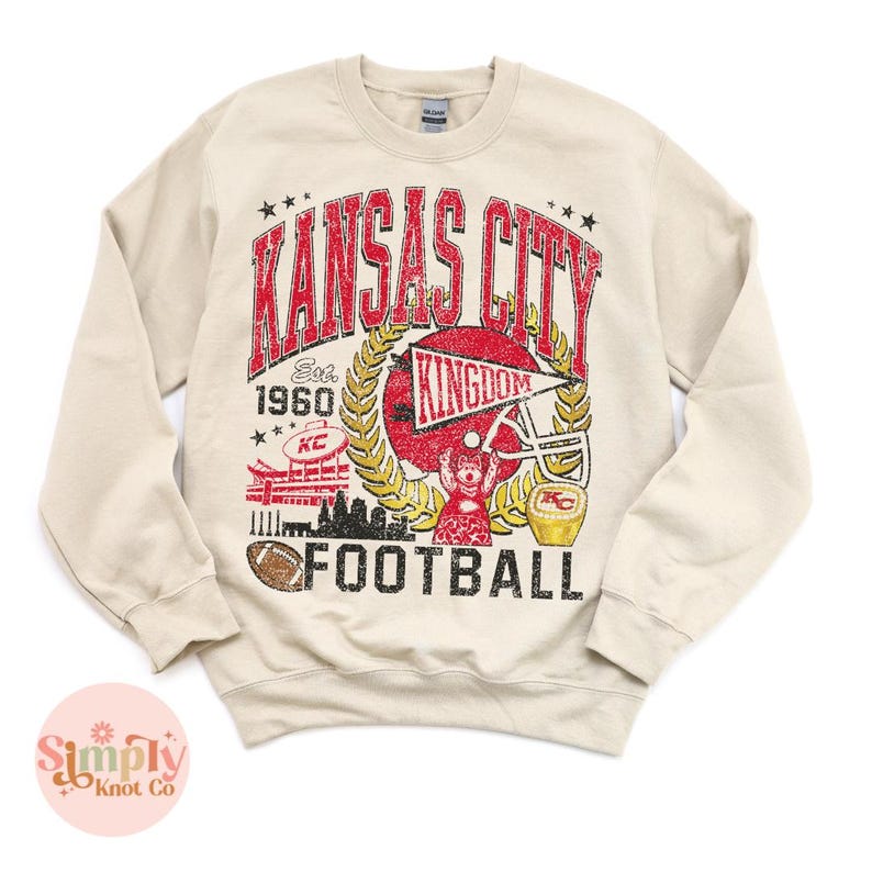Puede incluir: Sudadera beige con un dise&ntilde;o gr&aacute;fico desgastado. El dise&ntilde;o presenta las palabras "Kansas City Football" en rojo, con un casco de f&uacute;tbol, un bal&oacute;n de f&uacute;tbol y el texto "Est. 1960". La sudadera tiene cuello redondo y mangas largas.