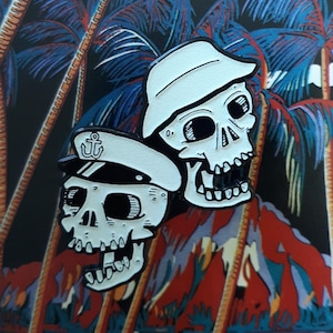 Puede incluir: Dos pines de esmalte blanco y negro que representan cráneos de dibujos animados con sombreros de marinero. El cráneo de la izquierda lleva un sombrero de marinero blanco y negro con un emblema de ancla. El cráneo de la derecha lleva un sombrero de marinero blanco. Los pines están sobre un fondo de palmeras.