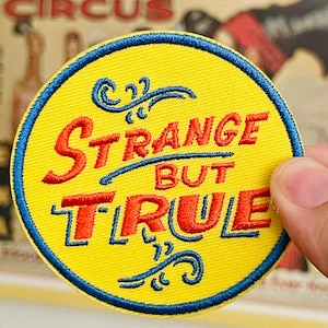 May include: A circular embroidered patch with a bright yellow background and a dark blue border. The text "STRANGE BUT TRUE" is stitched in red with a dark blue outline. Decorative blue flourishes are positioned above and below the text.