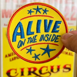 May include: A circular embroidered patch with a yellow background and a red border. The text ALIVE ON THE INSIDE is stitched in blue, flanked by three blue stars at the top and two blue stars at the bottom, all set against a yellow fabric base.