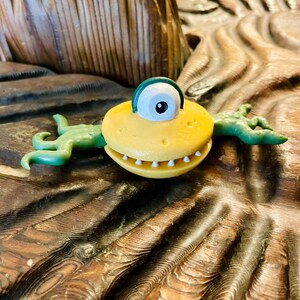May include: A yellow, circular creature figurine with a single large white eye featuring a black pupil and a dark green eyelid. The creature has a wide mouth with small white teeth and two green, multi-fingered arms extending from its sides.