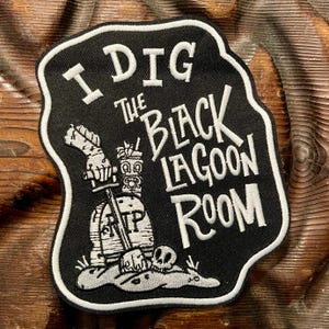 May include: A black embroidered patch with a white border featuring the text I DIG THE BLACK LAGOON ROOM. The design includes a hand holding a shovel stuck into a tombstone marked RIP, a tiki mug, and a skull resting on a mound of dirt.