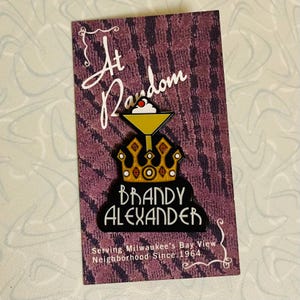 May include: A black and gold enamel pin with a crown and a cocktail glass with a cherry on top. The text "At Random Brandy Alexander" is on the pin. The text "Serving Milwaukee's Bay View Neighborhood Since 1964" is below the pin.