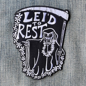 May include: Black and white embroidered patch featuring a skeleton figure in a hooded robe holding a scythe. The figure is wearing a flower lei and the text "LEID TO REST" is above the figure.