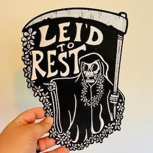 Peut inclure: Patch brodé noir et blanc représentant un squelette portant un collier de fleurs et tenant une faux. Le texte "Lei'd to Rest" est au-dessus du squelette.