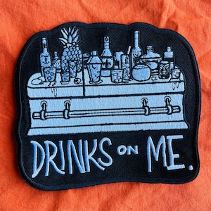May include: Black and white embroidered patch with a coffin and various bottles of alcohol on top. The text "DRINKS ON ME." is below the coffin.
