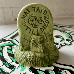 May include: A green ceramic tiki mug shaped like a tombstone. The tombstone has the words "Mai Tai'd Till Died" and a skull graphic. The base of the mug has a grass and flower design. The mug is a unique and spooky addition to any barware collection.