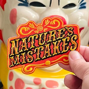 May include: A rectangular embroidered patch with a red background and a yellow border. The text "NATURE'S MISTAKES" is stitched in black with yellow highlights in a stylised font. Decorative yellow scrollwork accents the top and bottom edges.