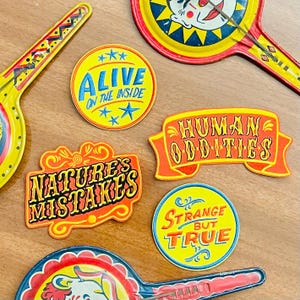 May include: A collection of colourful metal signs and circular badges featuring vintage-style typography. The items include text reading ALIVE ON THE INSIDE, HUMAN ODDITIES, NATURE'S MISTAKES, and STRANGE BUT TRUE, alongside two metal hand mirrors with clown graphics.