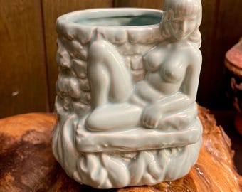 May include: A light blue ceramic planter with a reclining female figure. The planter has a textured, brick-like exterior and a detailed sculpture of a woman resting on a ledge. The overall design is artistic and decorative.