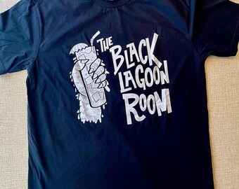 May include: A black short-sleeve t-shirt featuring a white graphic print of a scaly hand holding a tall glass with a straw and ice cubes. The text "THE BLACK LAGOON ROOM" is printed in large white letters next to the illustration on the front.