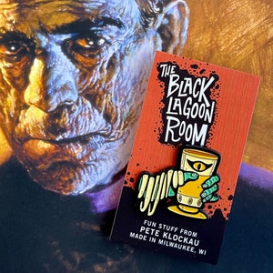 May include: A black and green enamel pin featuring a creature from the Black Lagoon holding a glass of beer. The pin says "The Black Lagoon Room" and "Fun stuff from Pete Klockau Made in Milwaukee, WI".
