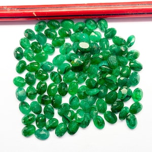 Può includere: Una collezione di gemme di smeraldo taglio ovale in varie tonalità di verde. Le gemme sono sparse su una superficie bianca, con una matita rossa visibile nella parte superiore dell'immagine. Gli smeraldi hanno un aspetto lucido e sfaccettato.