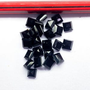 Puede incluir: Un conjunto de pequeñas gemas negras de corte cuadrado dispersas sobre una superficie blanca. Las gemas tienen una superficie pulida y reflectante. Un lápiz rojo y negro es visible en el fondo.